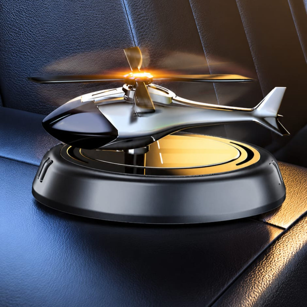 A miniature silver and black helicopter toy or model hovering above a circular black base on a dark leather surface, with illuminated golden rotor blades and a warm light reflection.