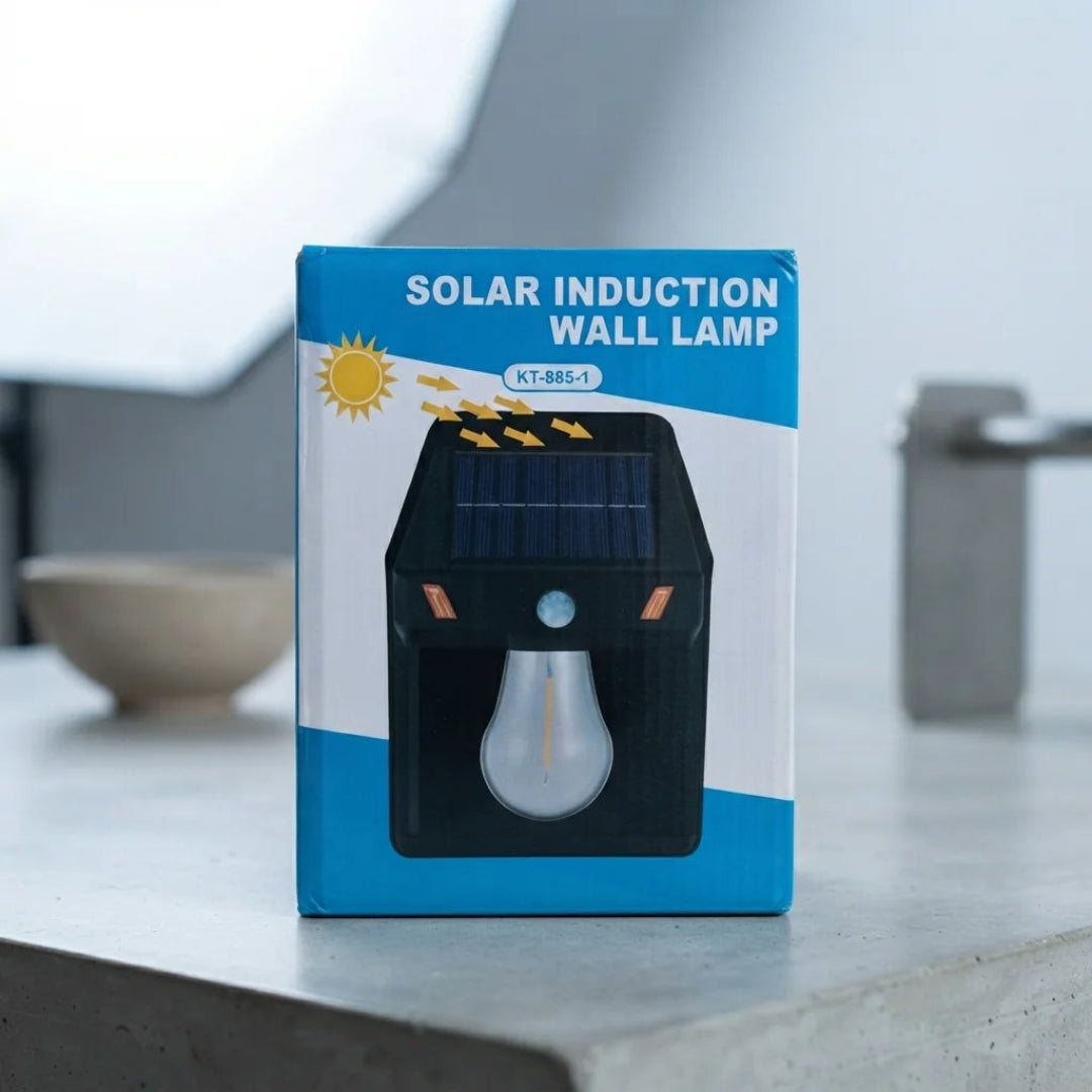A boxed solar induction wall lamp standing on a light gray countertop in a real indoor setting, with a bowl and faucet blurred in the background. The package is blue, white, and black with a printed solar panel and bulb illustration and the text 'SOLAR INDUCTION WALL LAMP'.