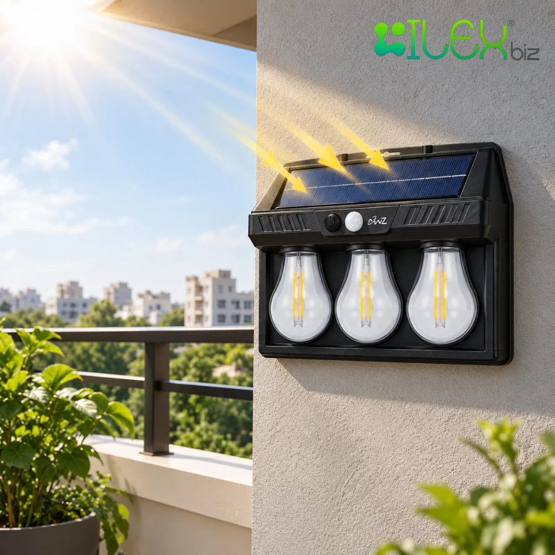 A black solar-powered outdoor light fixture mounted on a beige wall, featuring three clear bulb-shaped LED lights and a small solar panel on top. Yellow rays are illustrated shining onto the panel, and the background shows a balcony with plants and buildings under a blue sky.