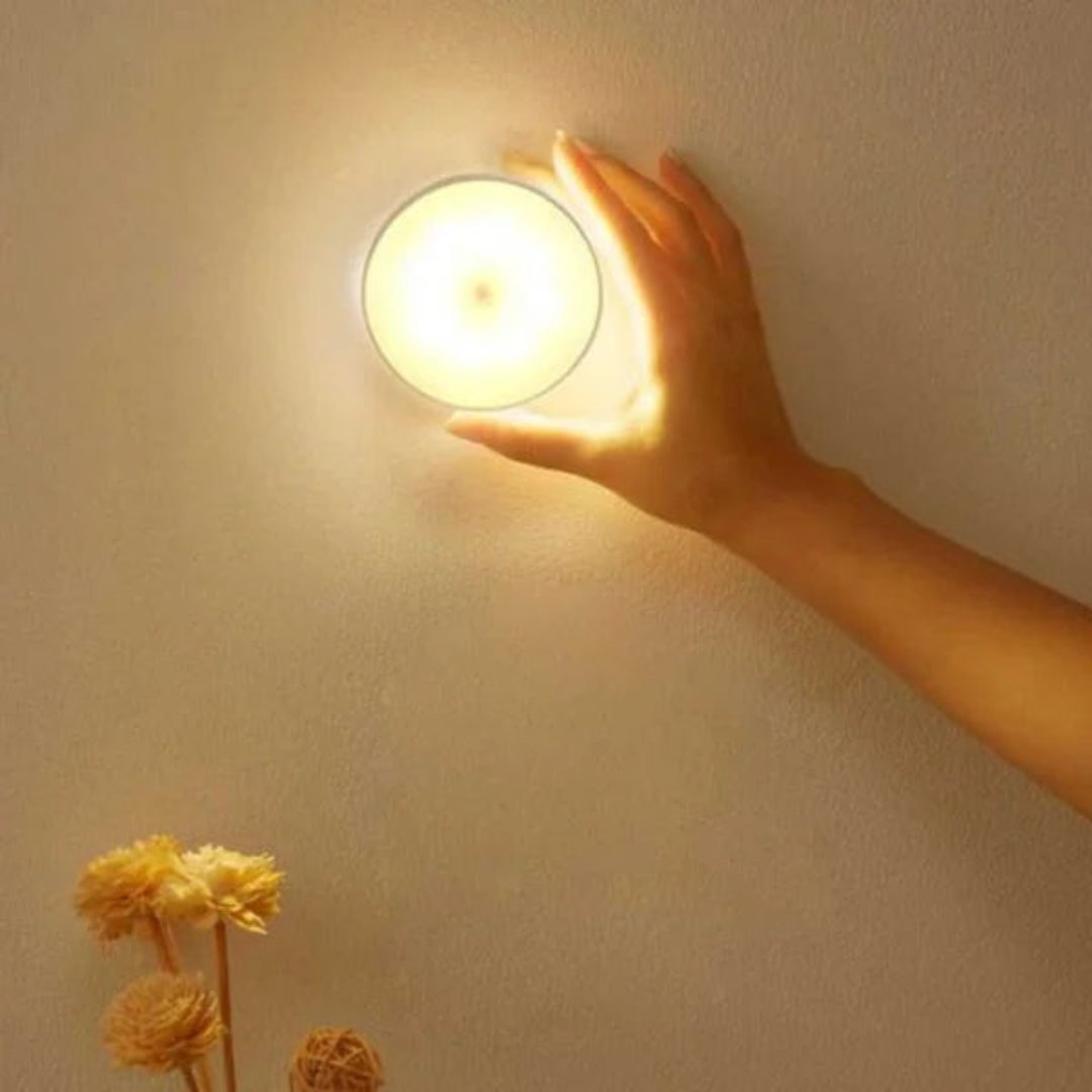 A hand touching a glowing round wall light mounted on a beige wall, with dried flowers in the lower left corner.