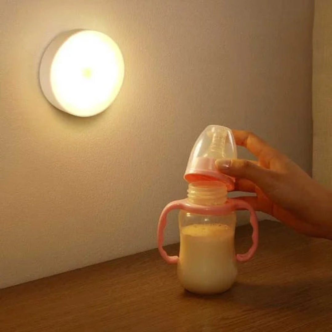 A hand holds a pink baby bottle with a clear cap and pink handle, partially filled with milk, while a glowing round wall light illuminates a beige wall above a wooden surface.