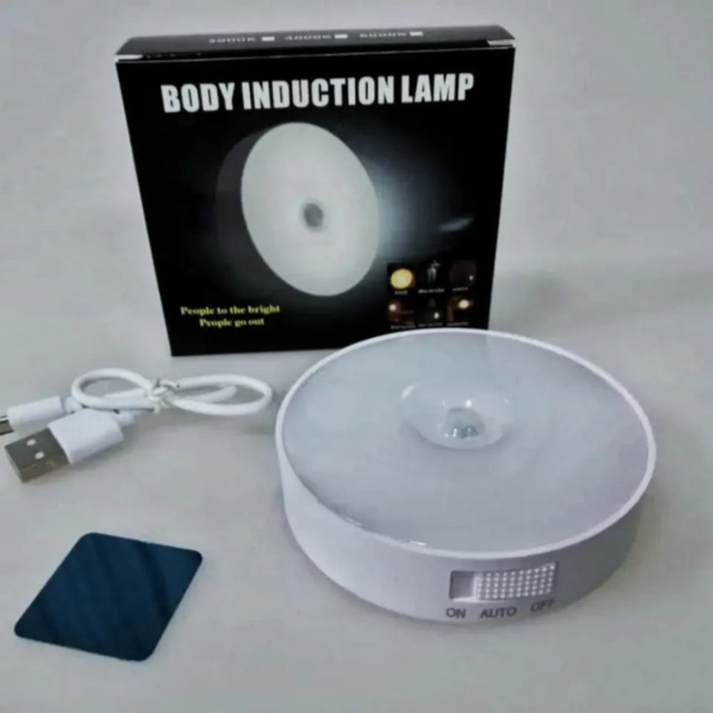 A white circular body induction lamp shown with its black retail box, a white USB charging cable, and a small dark blue square accessory on a light gray background.