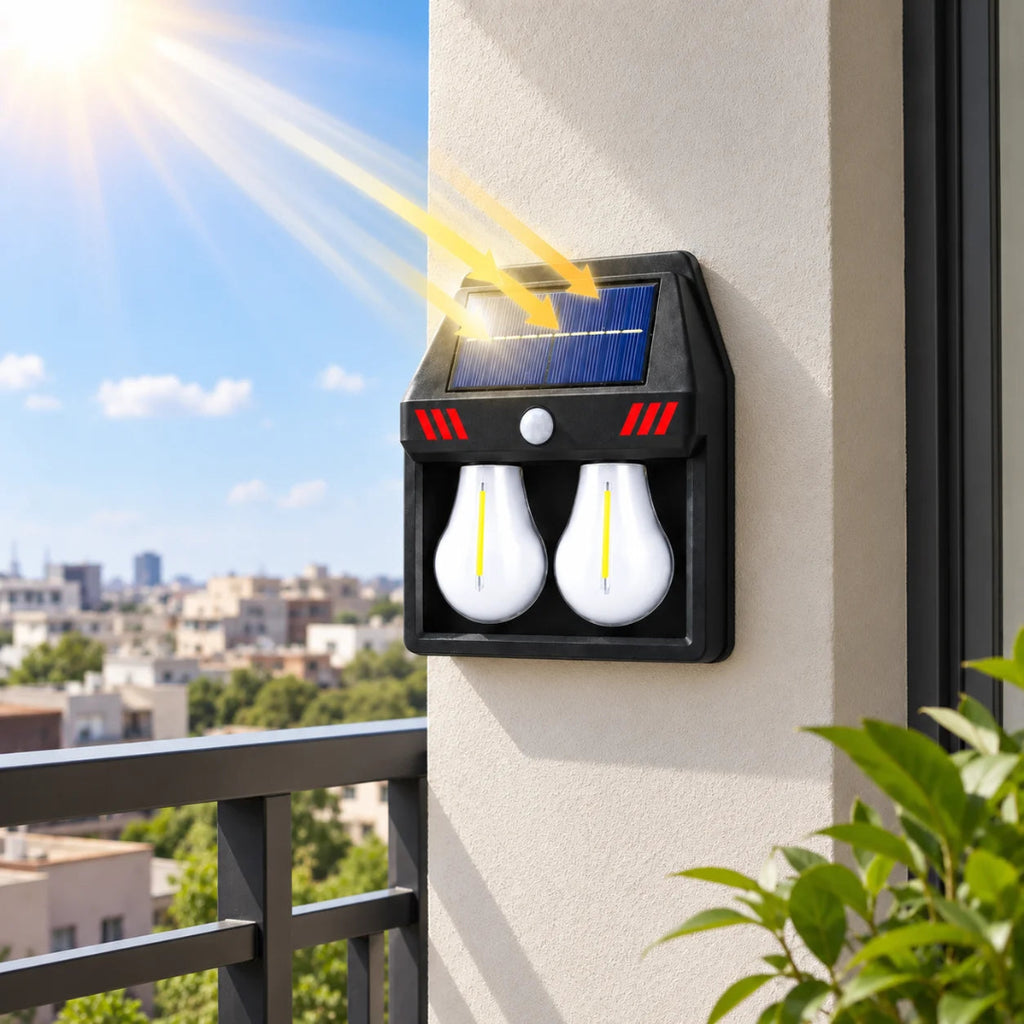 A black solar-powered dual-light outdoor wall sconce mounted on a beige exterior wall, with a visible solar panel, motion sensor, and two glowing white bulbs. Sun rays shine onto the solar panel, and a balcony railing with a city skyline is visible in the background.