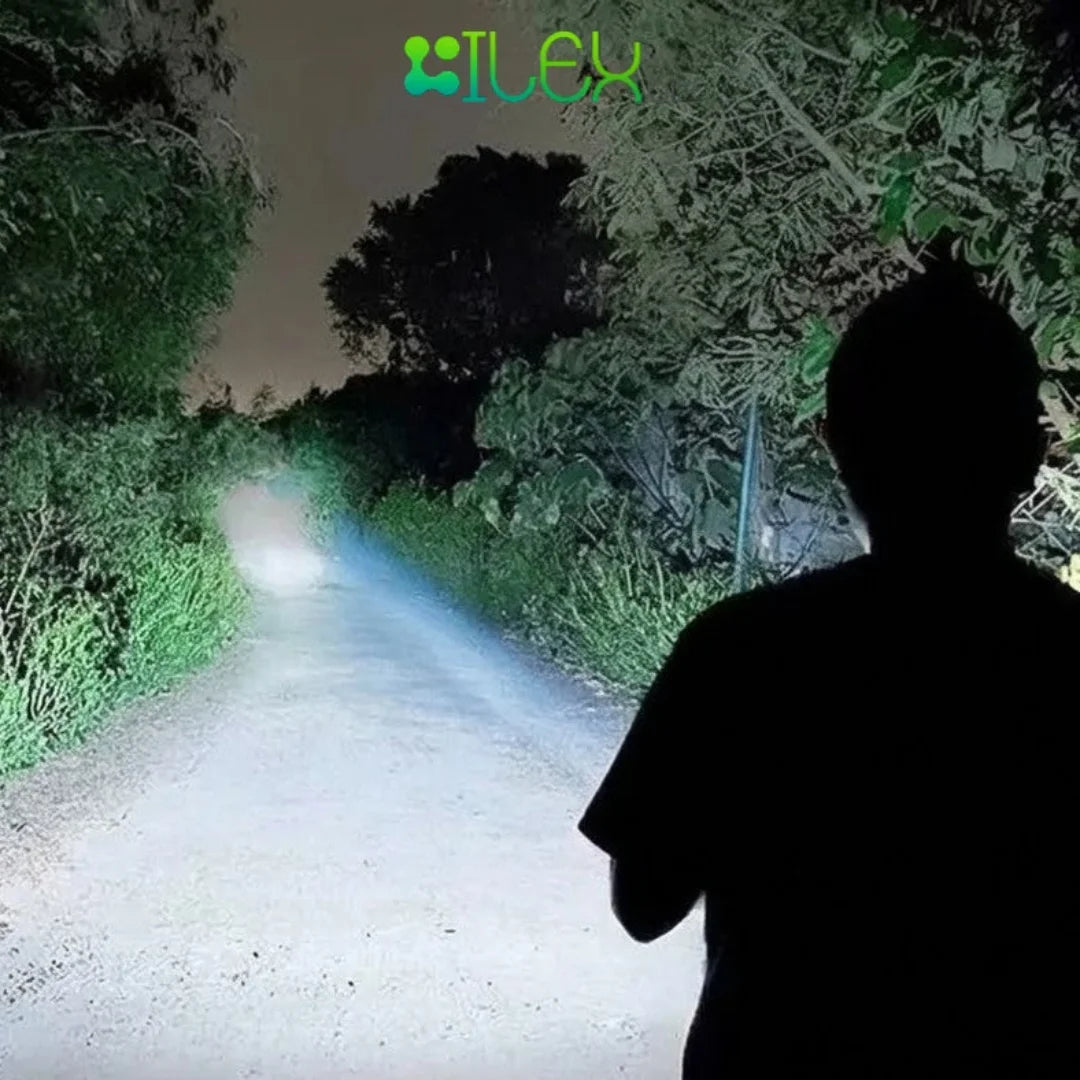 A person shown from behind in silhouette, standing on a dirt path at night with a bright LED flashlight beam illuminating the road ahead through dense green foliage. The brand name 'iLex' is visible at the top of the image.