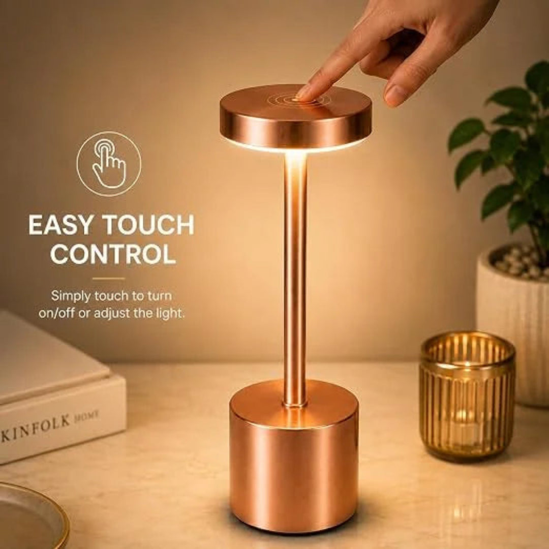 A copper-toned LED table lamp with a cylindrical base and stem, shown on a tabletop with a hand touching the top to demonstrate touch control.