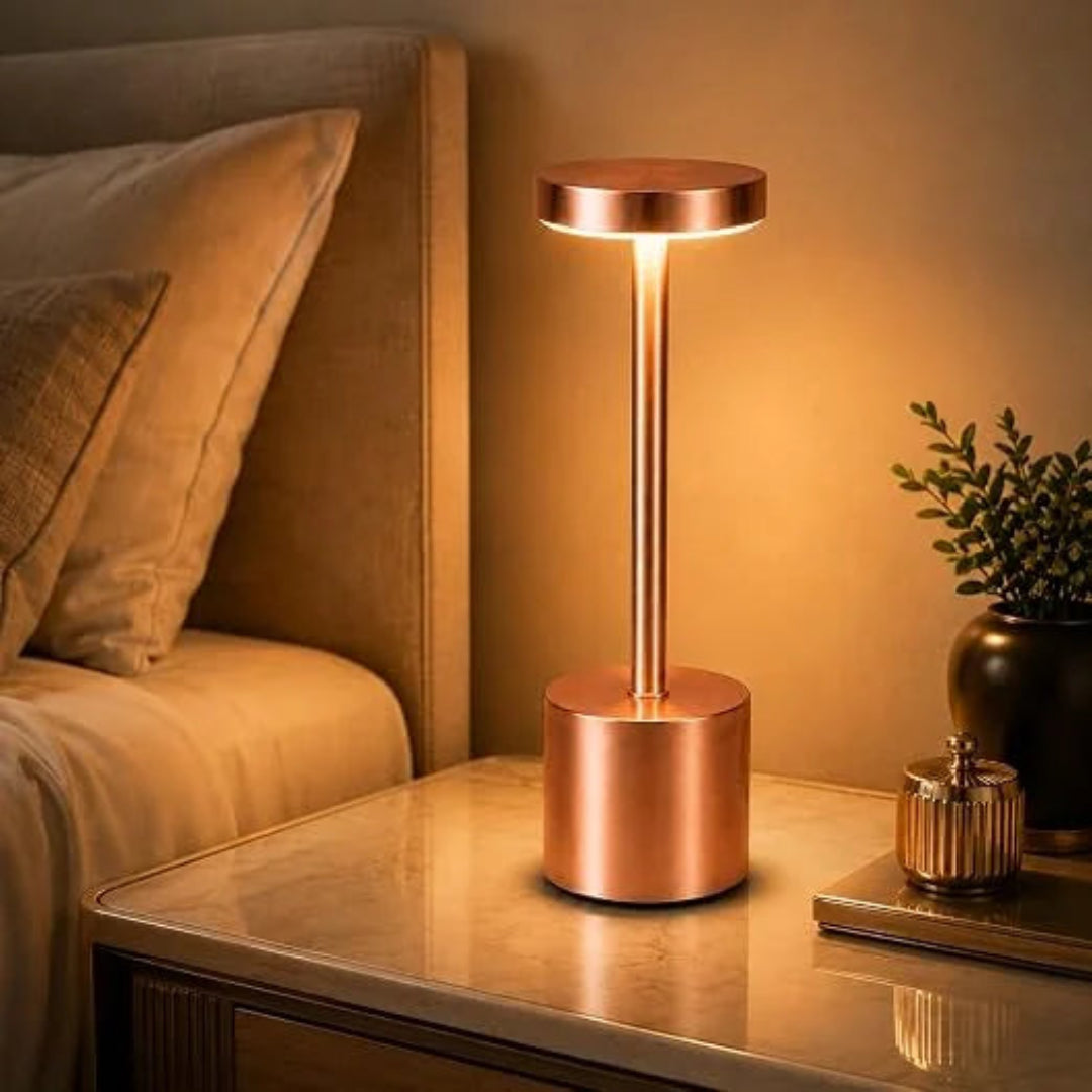 A modern table lamp with a cylindrical copper-finished base and matching round shade, illuminated with a warm glow on a bedside table next to a bed.