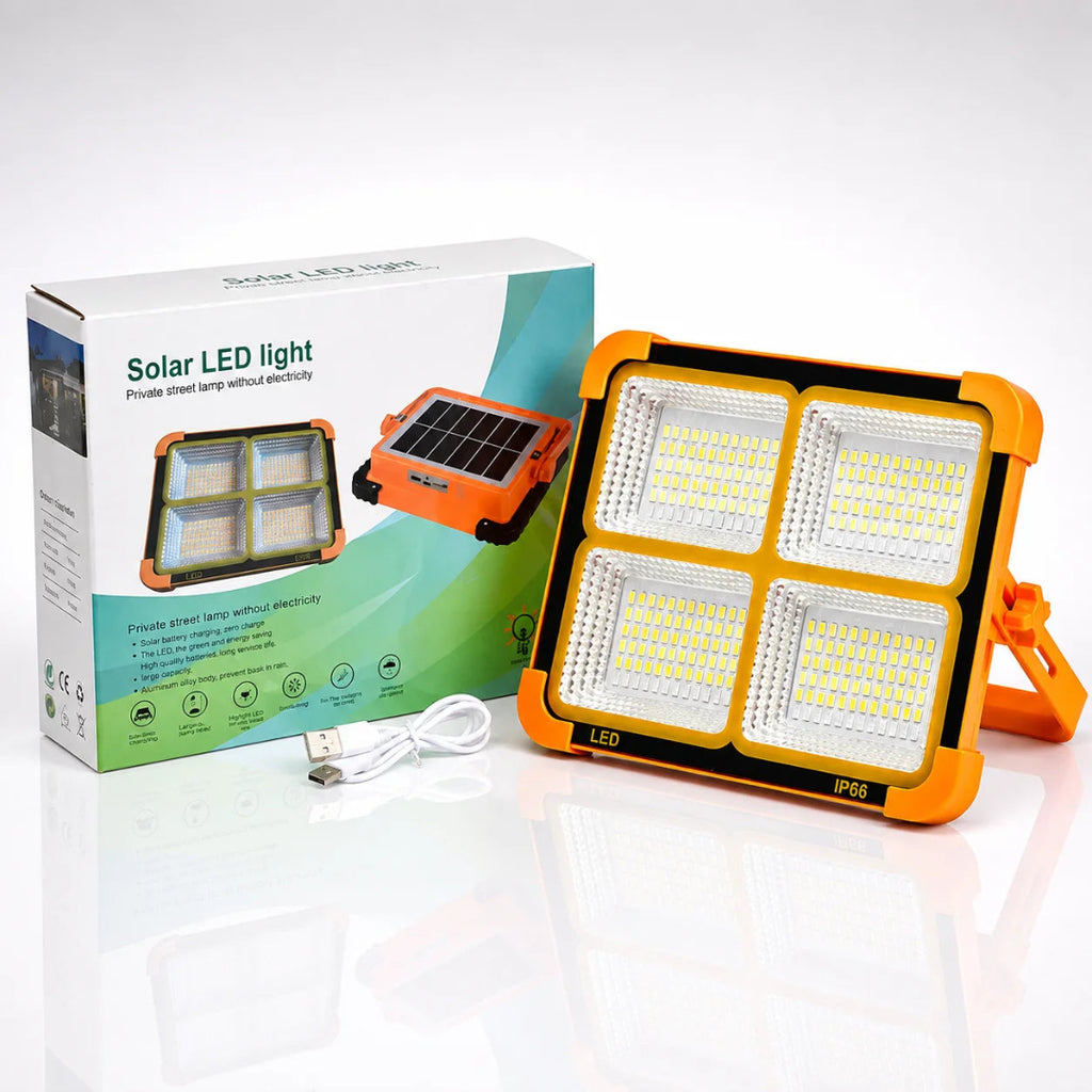 Solar LED light with a bright orange casing and four large white LED panels, shown next to its white and green retail box with a USB charging cable on a reflective white surface.