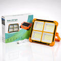 Solar LED light with a bright orange casing and four large white LED panels, shown next to its white and green retail box with a USB charging cable on a reflective white surface.