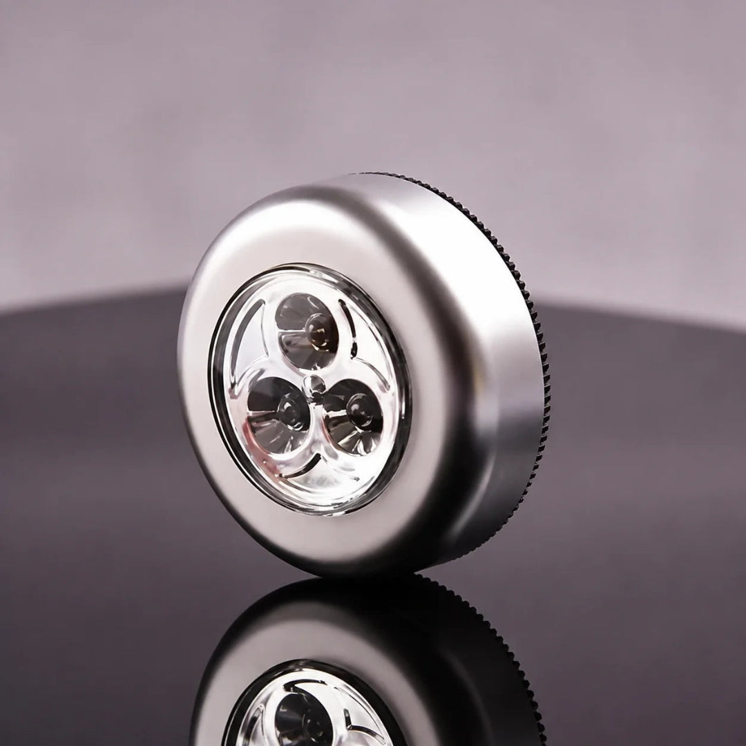 A round metallic silver device with a clear front lens showing three internal light elements, photographed on a dark reflective surface against a soft gray background.