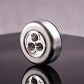 A round metallic silver device with a clear front lens showing three internal light elements, photographed on a dark reflective surface against a soft gray background.