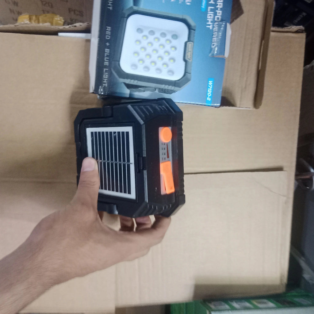 A hand holding a black and orange portable solar LED light in front of a cardboard box, with its retail packaging box visible in the background.