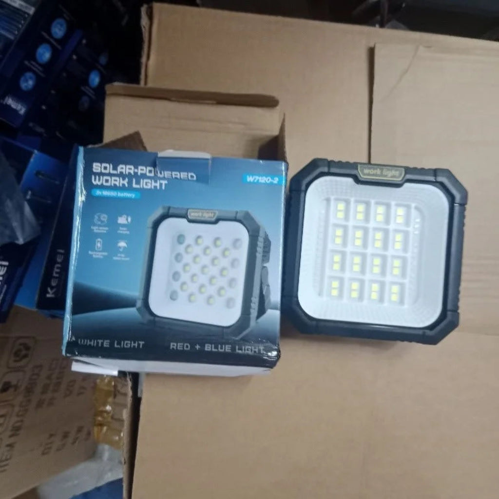 A solar-powered work light shown next to its retail box on a cardboard shipping box. The product is a square rugged LED work light with a black protective frame and a white face containing multiple LED chips.