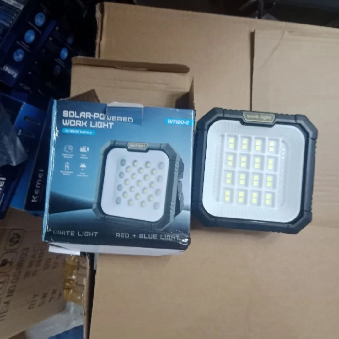 A solar-powered work light shown next to its retail box on a cardboard shipping box. The product is a square rugged LED work light with a black protective frame and a white face containing multiple LED chips.