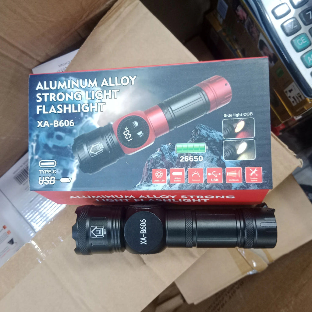 A black aluminum flashlight with a red end cap placed in front of its retail box, which reads 'ALUMINUM ALLOY STRONGLIGHT FLASHLIGHT' and model 'XA-B606'. The scene is photographed inside a cardboard box with a calculator visible in the background.